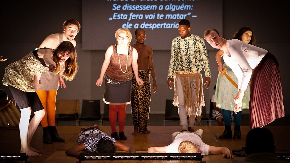 Participants on a stage. Two people are lying stretched out on the floor. There are eight other people standing behind them. In the background there is a screen with words on it.