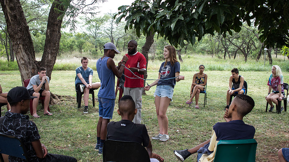 Participants sitting in a circle, with two of them standing opposite each other in the middle of the circle. They are tied together by a rope around their wrists. A third person is standing next to them.