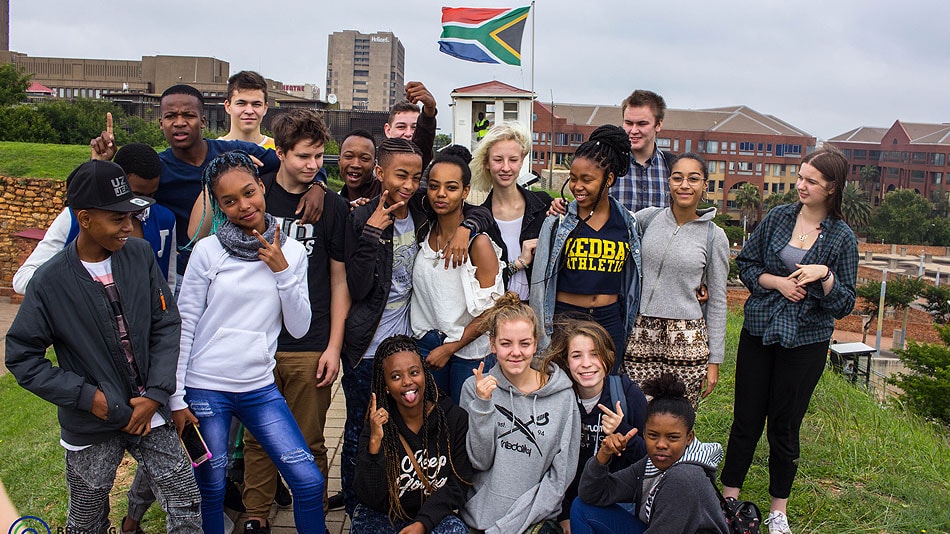 Gruppenfoto einer teilnehmenden Jugendgruppe auf einer Wiese stehend. Im Hintergrund sind Gebäude und die südafrikanische Fahne auf einem Fahnenmast.