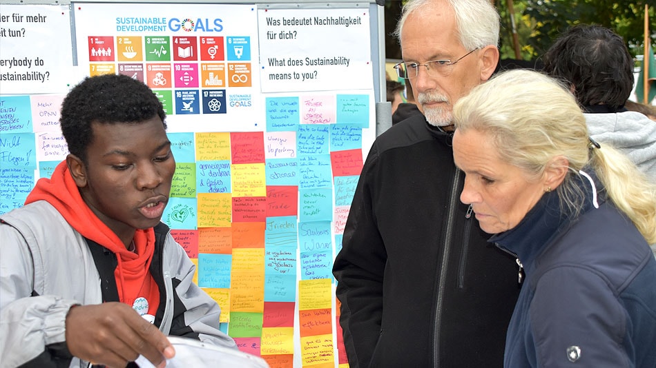 Ein Teilnehmender steht an einem Ausstellungsstand und erklärt einem Mann und einer Frau etwas. Im Hintergrund hängen Plakate mit den Entwicklungszielen und der Frage „Was bedeutet Nachhaltigkeit für dich“?