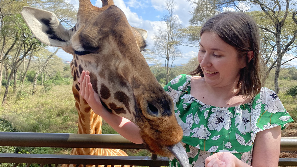Une personne perçue comme étant une femme se tient devant un enclos de girafes et caresse la tête de l'une d'entre elles d'un air amusé. La girafe tire la langue.