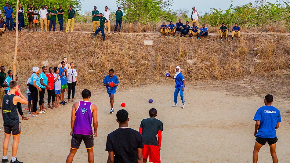 La photo montre un entraînement de volley-ball. Plusieurs jeunes se trouvent sur le terrain de sable. Un jeune court avec le ballon, une autre se prépare à lancer. D'autres personnes se tiennent debout ou assises sur le bord et sur le chemin un peu plus h