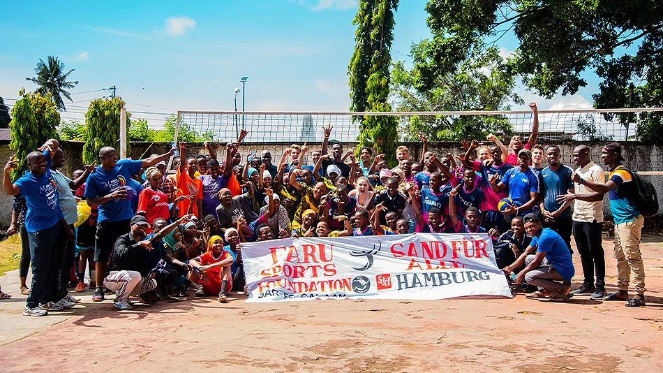 This photo shows a large group of people on a volleyball court. They have assembled in front of the net and some of them are holding up a banner with the names of the two exchange partners on it: FARU Sports Foundation Dar es Salaam and Sand für Alle Hamb