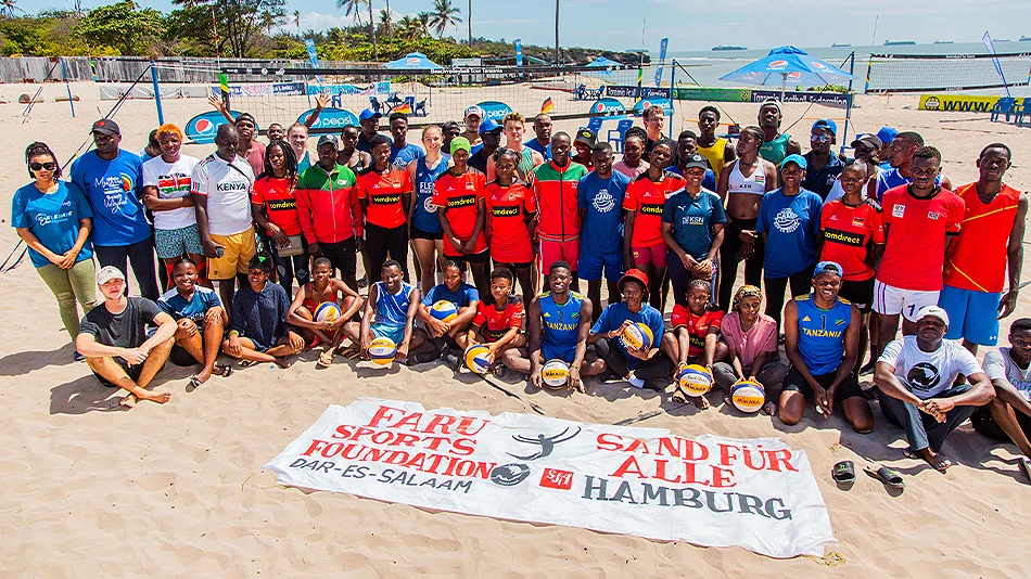 Für dieses Bild stellen sich die Sportlerinnen und Sportler auf dem Volleyballgelände auf. Im Vordergrund liegt auf dem Sand das Banner der beiden Kooperationspartner FARU Sports Foundation Dar-Es-Salaam und Sand für Alle Hamburg.