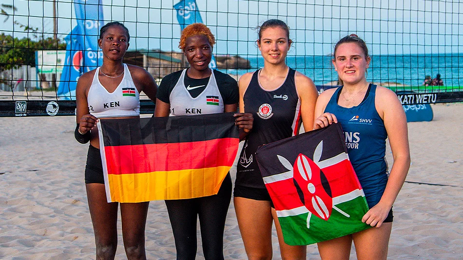 Dieses Foto zeigt vier Personen vor dem Netz eines Beachvolleyball-Feldes. Links stehen zwei Volleyballerinnen aus Kenia, die eine deutsche Flagge präsentieren, und rechts zwei Volleyballerinnen aus Deutschland mit einer Kenia-Fahne.