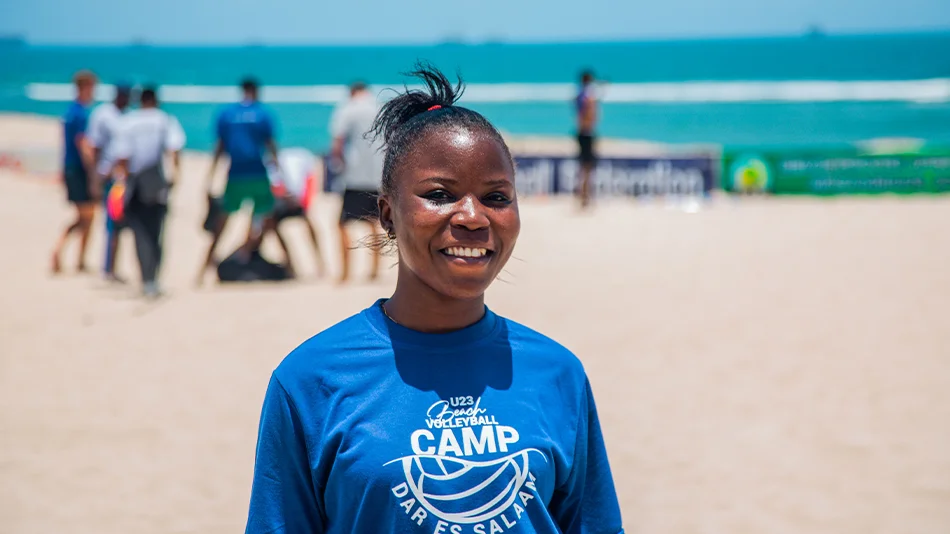 Dieses Foto zeigt die Teilnehmerin Victoria John Mwenele lächelnd vor den Volleyballfeldern am tansanischen Strand.