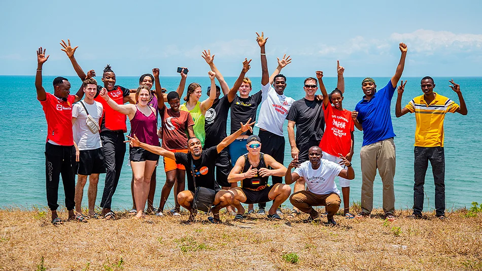 The photo shows 17 of the youth exchange participants, who have assembled for a group photo. They are standing on dry grass, with the sea in the background, and joyfully waving their arms in the air.