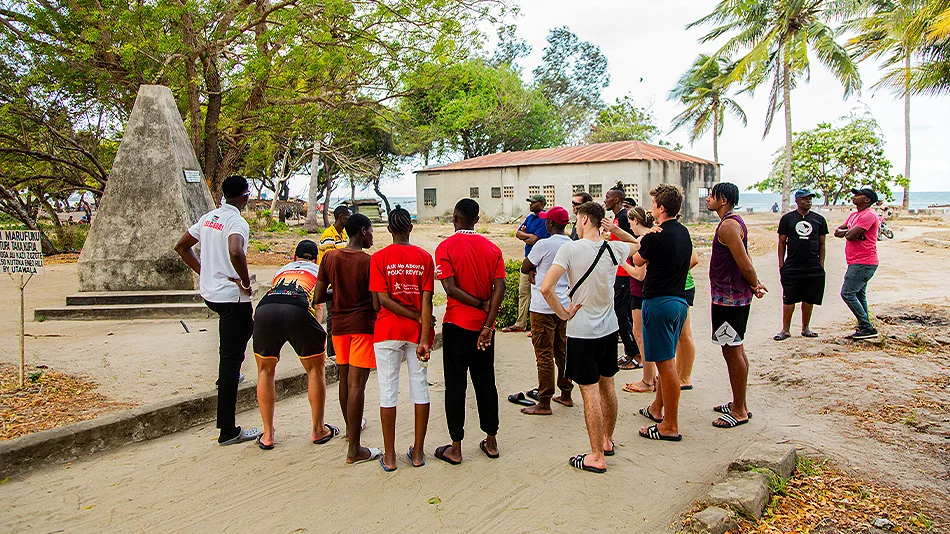 La photo montre au moins 17 personnes qui se tiennent autour d'un monument en pierre en forme de cône. Il se trouve à Bagamoyo, ancienne capitale de la Tanzanie à l'époque du colonialisme. Les jeunes semblent écouter un guide.