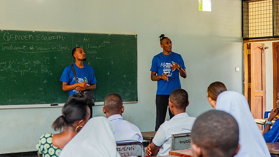 The photo shows two Tanzanian men, standing in front of a blackboard in a classroom and talking to seated people. The words “GENDER EQUALITY” can be seen in capitals on the blackboard.