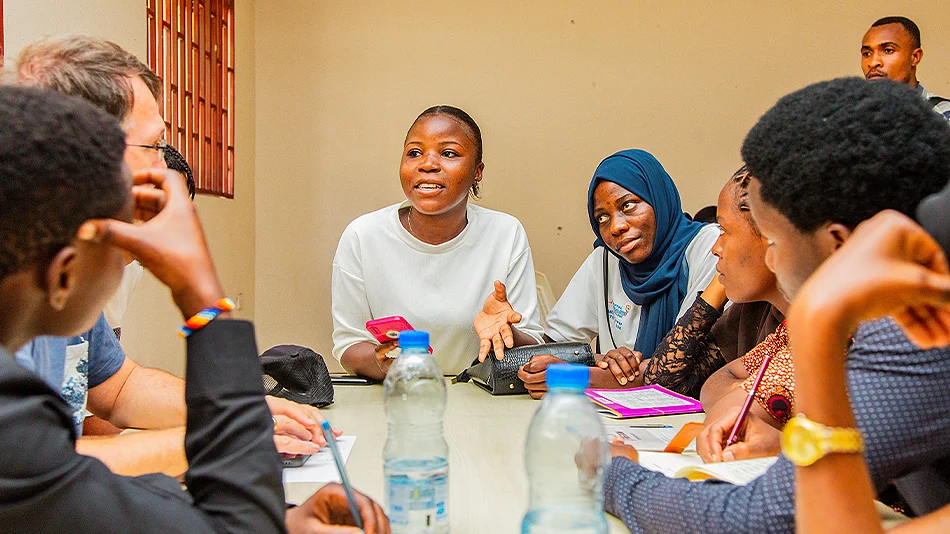 Cette photo montre au moins 7 participant·e·s à la rencontre de jeunes autour d'une table dans une salle de l'université de Kampala. Une personne parle, les autres l'écoutent attentivement.