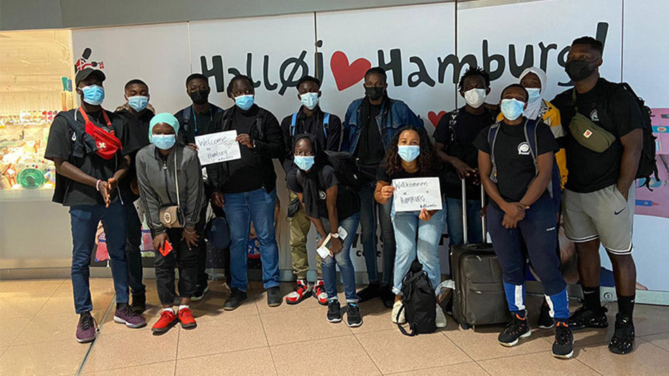 Bei ihrer Ankunft am Flughafen in Hamburg stehen dreizehn junge Schwarze Personen mit Maske vor einer Wand, auf der "Halloj Herz Hamburg!" steht. Die Personen sind Teilnehmende der Partnerorganisation DUNK (Develop Unity Nurturing Knowledge) aus Accra.
