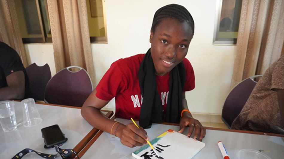 A young participant from partner organisation DUNK is smiling at the camera at a workshop in Accra. She is writing characters in black paint on a white tile or canvas.