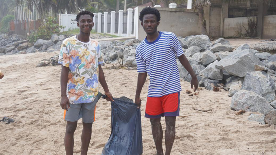 Zwei junge, männliche Teilnehmende der Partnerorganisation DUNK halten gemeinsam eine dunkle Mülltüte am Strand von Wakanda in Accra.