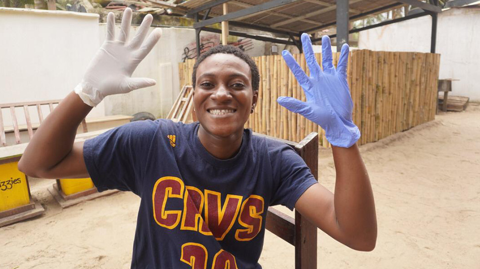 Une jeune participante de l'organisation partenaire DUNK sourit et montre ses mains, recouvertes de gants bleus et blancs, à la caméra. Derrière elle, des structures en bois et en pierre difficiles à définir d'une usine de recyclage à Accra.
