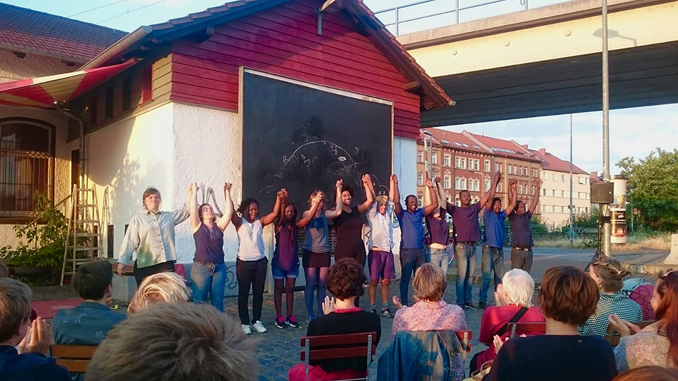 The participants are standing in a row, hand in hand, ready to take a bow before an audience. The audience is applauding. The photograph was taken in an urban setting with terraced housing in the background.