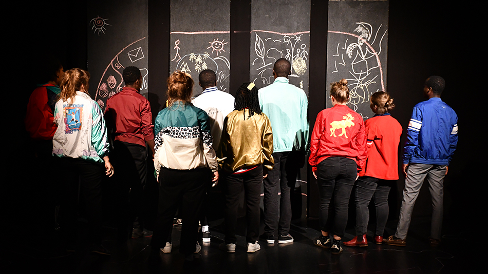 Le groupe se tient sur la scène. Un dessin à la craie se trouve sur le mur noir. On aperçoit les participant·e·s de dos. Ils portent des pantalons de couleur sombre et des vestes colorées.