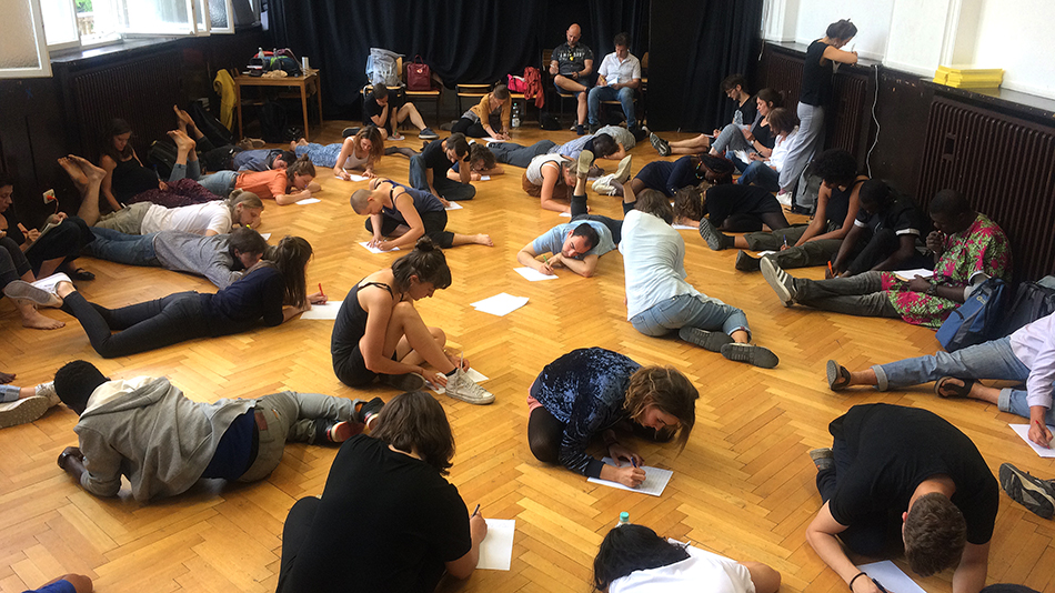 Lots of participants are lying or sitting on the floor, each writing on their own piece of white paper.