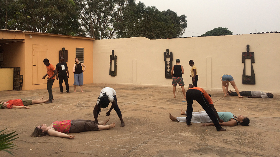 Some participants are lying flat on the ground in the rehearsal for their play. Other participants are bending over the people on the ground. An orange building and trees can be seen in the background.