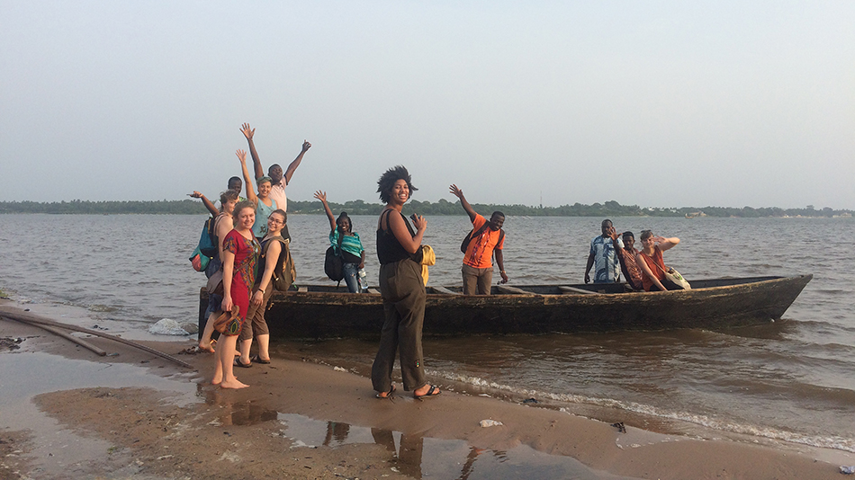 Participants on a trip to the seaside. Some people are sitting in a boat, others are still standing on the beach. They are smiling at the camera and some of them have their arms raised in the air.