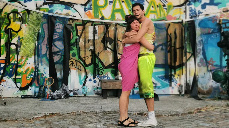 Une jeune femme et un jeune homme se tiennent dans les bras. Ils sont vêtus de couleurs vives et se tiennent devant le mur d'un immeuble couvert de graffitis.