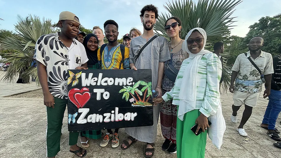 Eine Gruppe von jungen Menschen hält ein Schild mit der Aufschrift „Welcome to Zanzibar“. Im Hintergrund sind Palmen zu sehen.