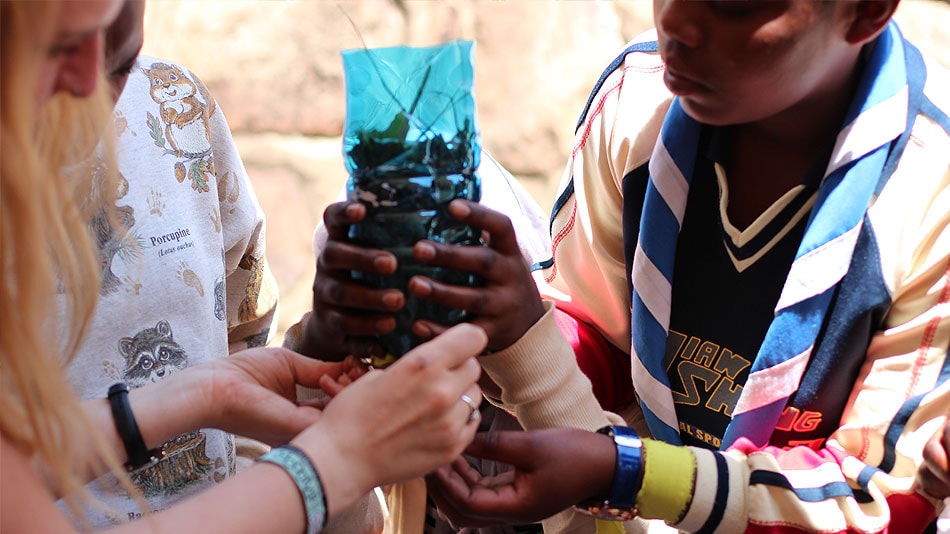 Three participants hold up a filter they built using twigs and a plastic bottle. One of them is wearing the scarf typically worn by scouts and guides.