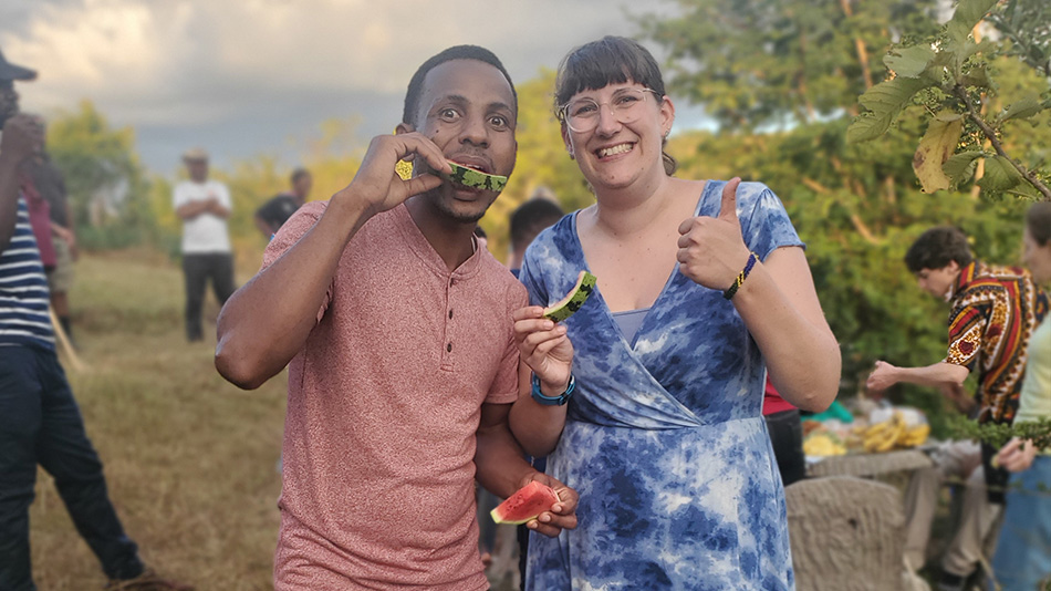 The photo shows Carolin and a colleague. They are eating watermelon and smiling at the camera. Carolin is giving a thumbs-up with her left hand.