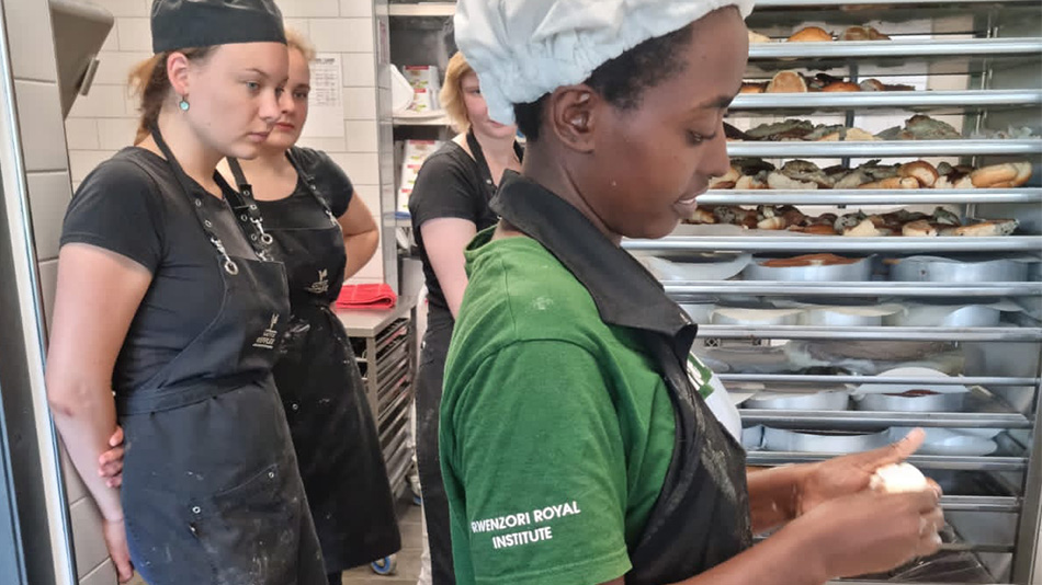 The foreground of the picture shows Rebecca, a young professional from Uganda. She is wearing an apron and a baker’s cap and is working with dough. In the background, three of her colleagues from Kaffee Wippler are watching.