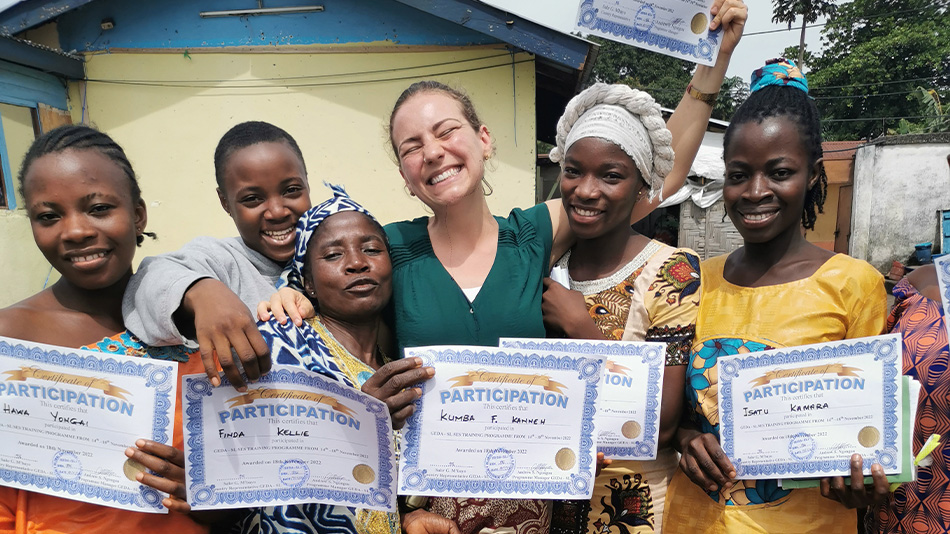 The photo shows a group of six young people who read as women, in front of a building. They are holding participation certificates up to the camera. Some of them have their arms around each other and are smiling. Marlena can be seen in the middle.