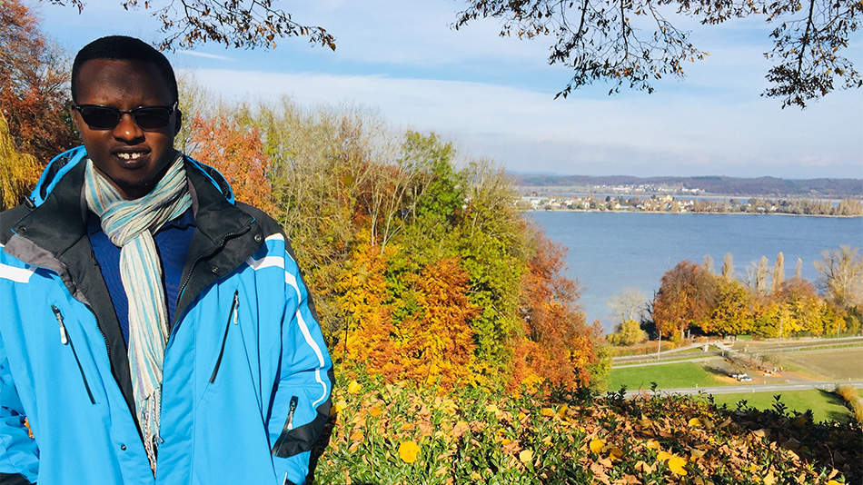 The photo shows Ole on the left half of the picture. He is wearing a blue winter jacket and is standing on a viewpoint. The right half of the picture shows an autumnal landscape and the Rhine.