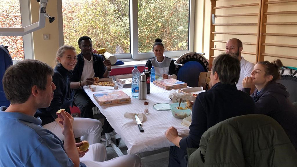 La photo montre un groupe de personnes assises autour d'une table. Certaines sourient à la caméra, d'autres sont en train de manger. L'une d'entre elles est Ole Lerug.