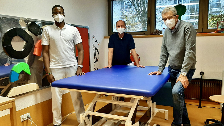 Trois personnes se tiennent dans une pièce autour d'une table de traitement à hauteur réglable. À côté de Ole se tiennent Stefan Daub, chef du service de kinésithérapie (au milieu) et le Dr. Willy Zink, expert du SES (à droite). Ils portent tous un masque
