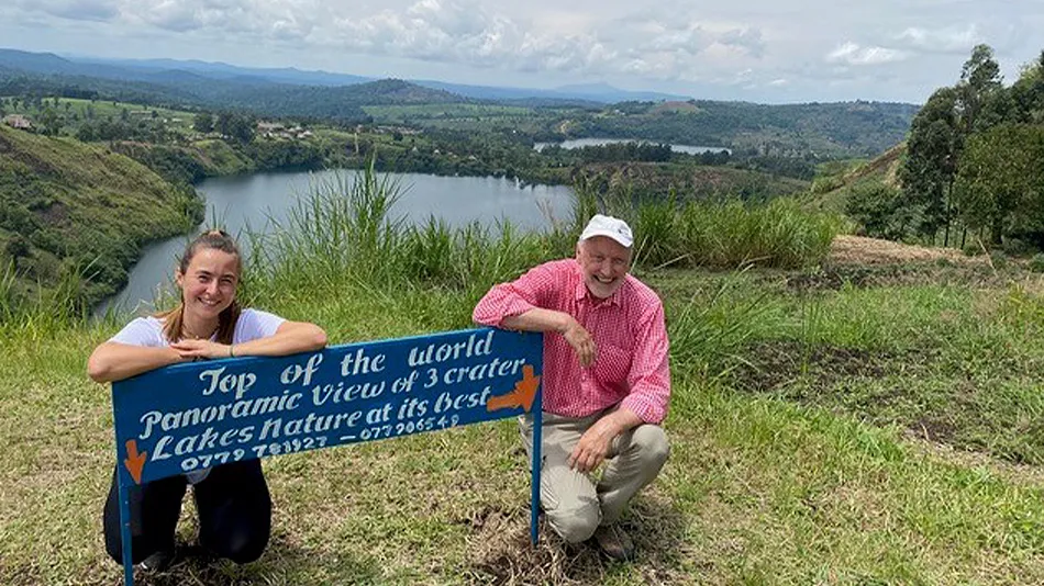 La photo montre Anna et Klaus. Ils/elles se trouvent tou·te·s les deux à un point de vue avec un lac derrière eux. Ils/elles sont agenouillé·e·s derrière un panneau bleu portant l'inscription « Top of the world » et sourient à la caméra.