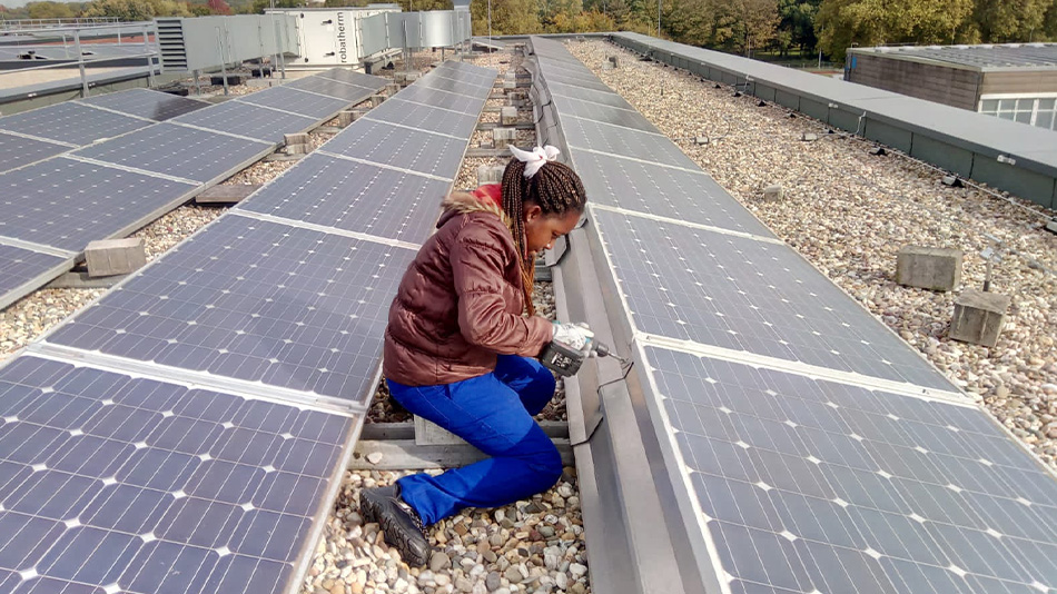 Erica is kneeling on a roof with many solar panels. Her gaze is directed at her hands, because she is using a cordless screwdriver.