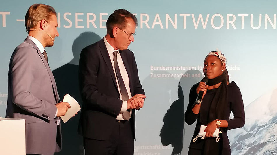 The former Minister Dr. Gerd Müller of the Ministry for Economic Cooperation and Development (BMZ) and another male person observe Erica who is holding a microphone and talks to them.