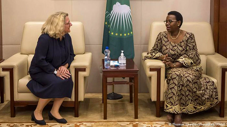 Zwei Frauen sitzen einander zugewandt und unterhalten sich. Zwischen beiden steht ein kleiner Tisch mit einer Wasserflasche und Desinfektionsmittel. Im Hintergrund hängt die dunkelgrüne Flagge der Afrikanischen Union mit gelben Sternen.