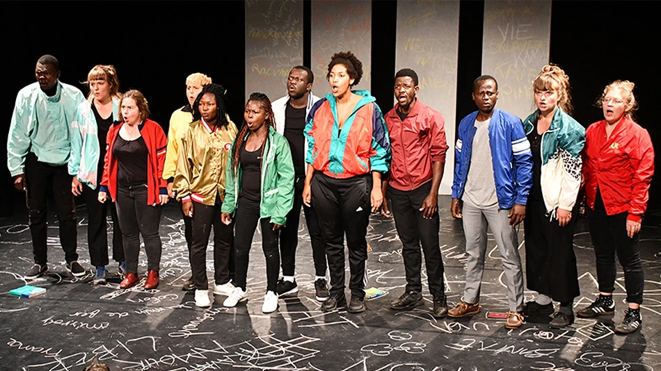 A group of 12 young people, some white, some BiPoC, are standing on a stage with white letters on a black floor. They are standing next to each other, looking out at the audience with their mouths open as they perform the play.