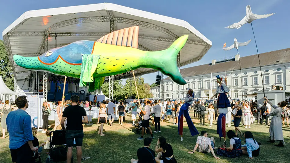 Das Bild zeigt viele Menschen vor einer Bühne im Park des Schlosses Bellevue. Menschen tragen einen großen bunten Fisch aus Pappmachée.