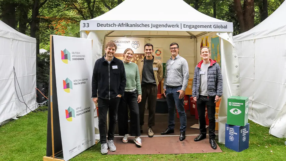 Fünf Personen stehen vor dem Zelt des DAJW auf dem Bürgerfest des Bundespräsidenten mit der Aufschrift „Deutsch-Afrikanisches Jugendwerk, Engagement Global“. Alle Personen gehören zum Team vom DAJW und lächeln in die Kamera. Hinter ihnen hängen Plakate.