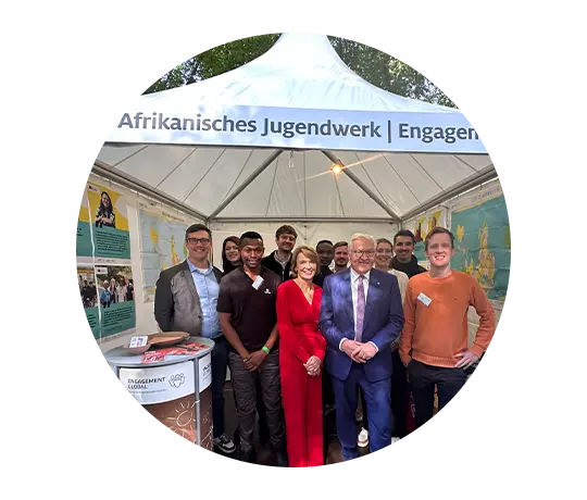 Das Bild zeigt eine Gruppe von Menschen unter einem weißen Zelt mit der Aufschrift „Deutsch-Afrikanisches Jugendwerk, Engagement Global“. Die zwei Personen vorne in der Mitte sind Bundespräsident Frank-Walter Steinmeier und seine Frau Elke Büdenbender.