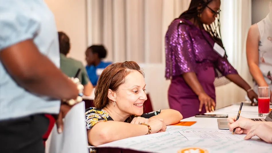 La photo montre une participante qui sourit. Elle regarde comment un autre participant écrit quelque chose sur un bout de papier.