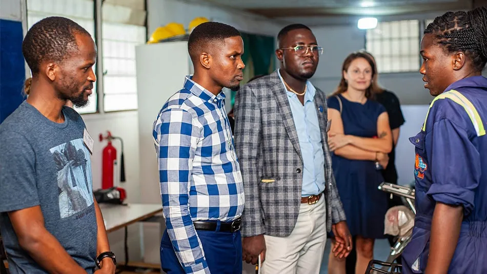 Das Bild zeigt eine Gruppe von Teilnehmenden, die einer Mitarbeiterin von Solar Taxi zuhören. Sie stehen in einer Werkstatt des Unternehmens.