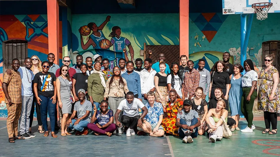 Das Bild zeigt eine große Gruppe auf dem Geländer von DUNK Grassroots in Jamestown. Die Gruppe besteht aus Workshopteilnehmenden, Mitgliedern von DUNK und von den Rising Lions.